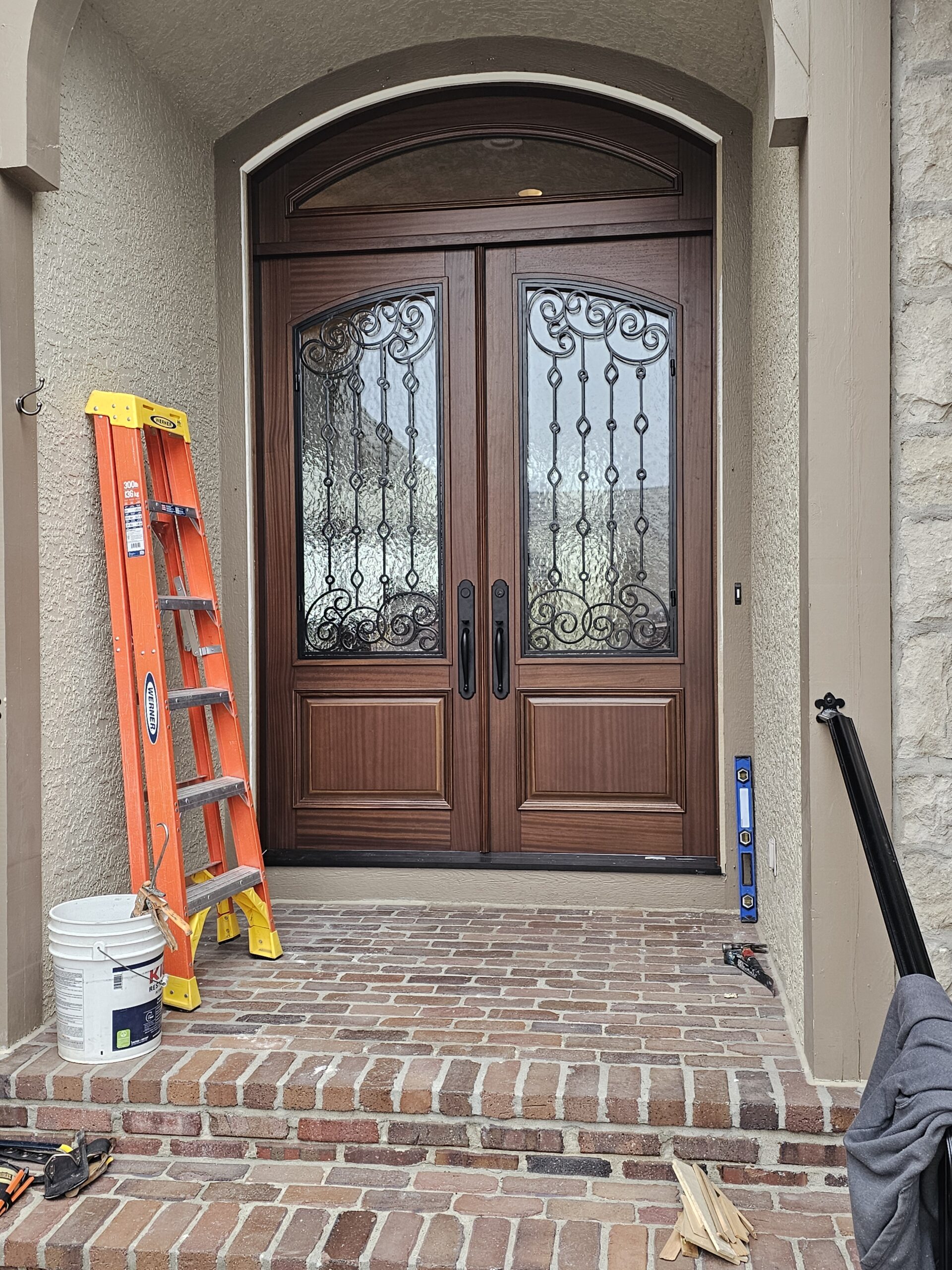 GlenviewDoor-KCM A decorative wood double front door with stone walls. On the left is an orange and yellow ladder leaning on the wall. Next to the ladder is a white bucket.