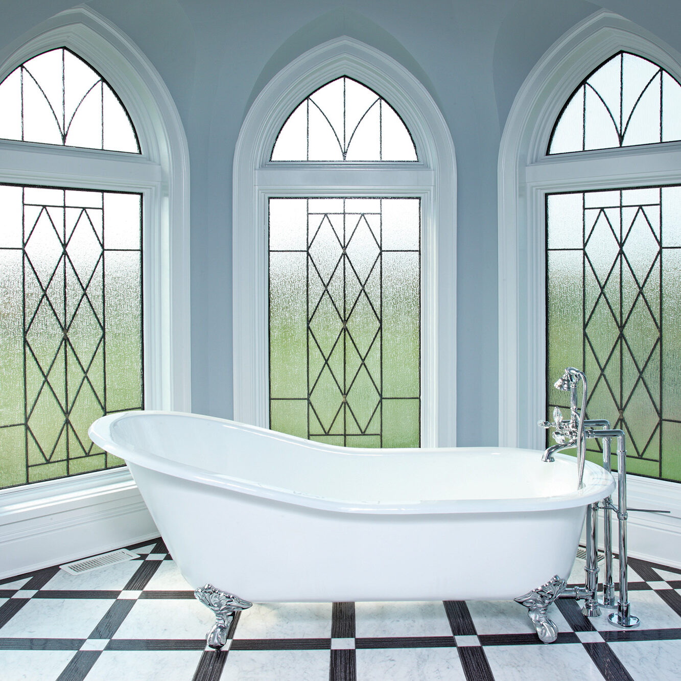 A white clawfoot bathtub sits in a room with three tall, arched windows. The walls are pale blue, and the floor has a checkerboard pattern.