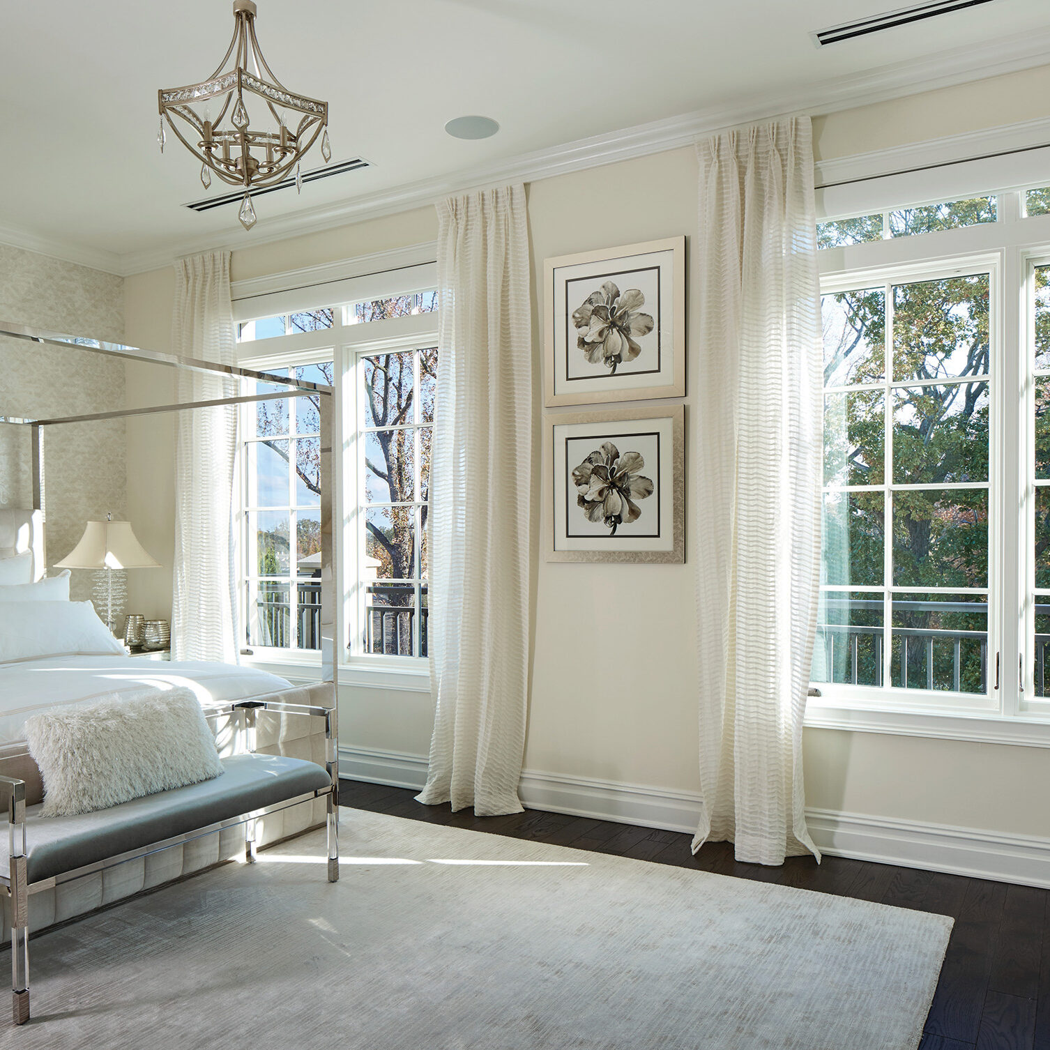Bright bedroom with a white canopy bed, gray bench, framed flower art, and large windows.