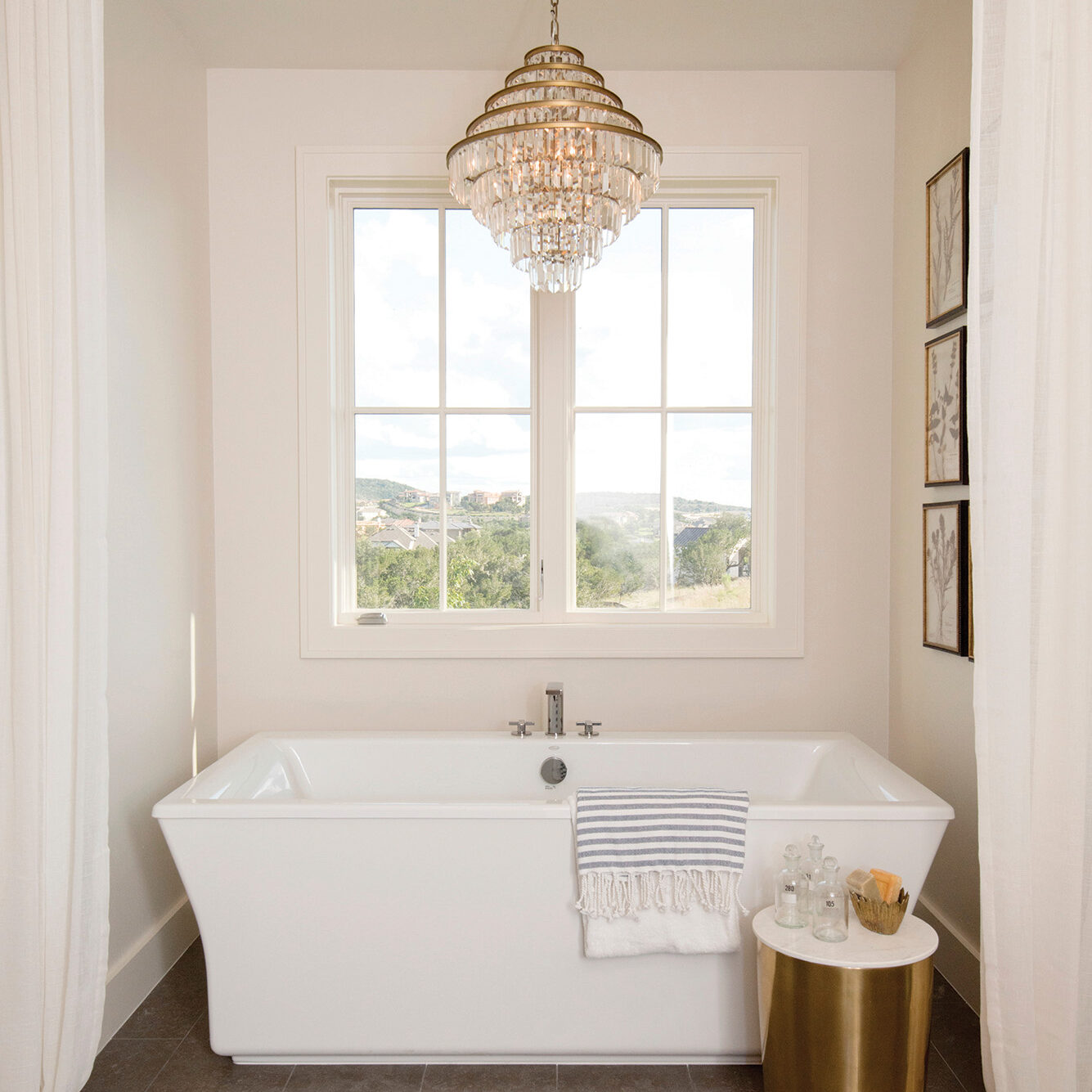 A stylish bathroom with a white bathtub, crystal chandelier, and a gold side table.