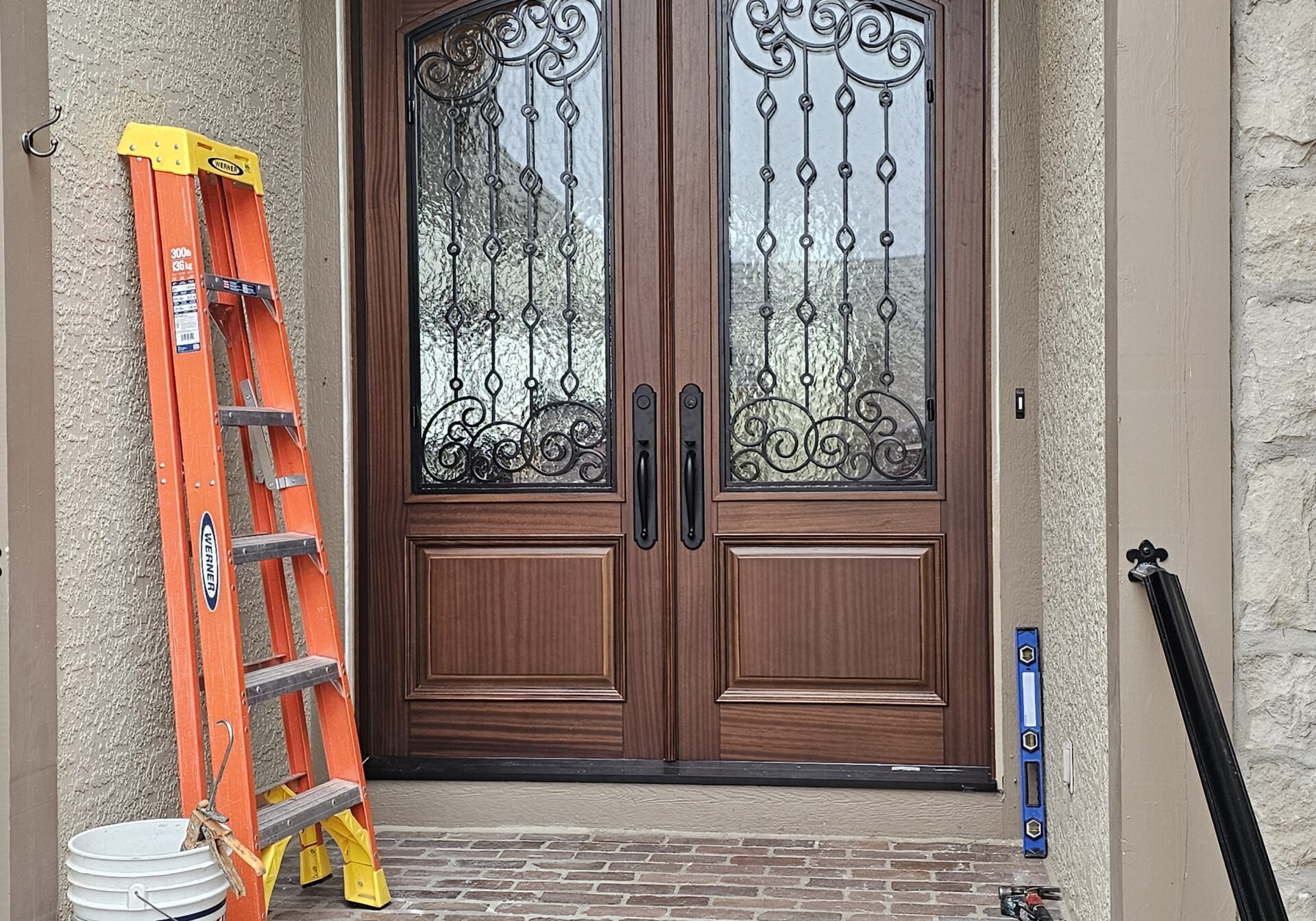 GlenviewDoor-KCM A decorative wood double front door with stone walls. On the left is an orange and yellow ladder leaning on the wall. Next to the ladder is a white bucket.