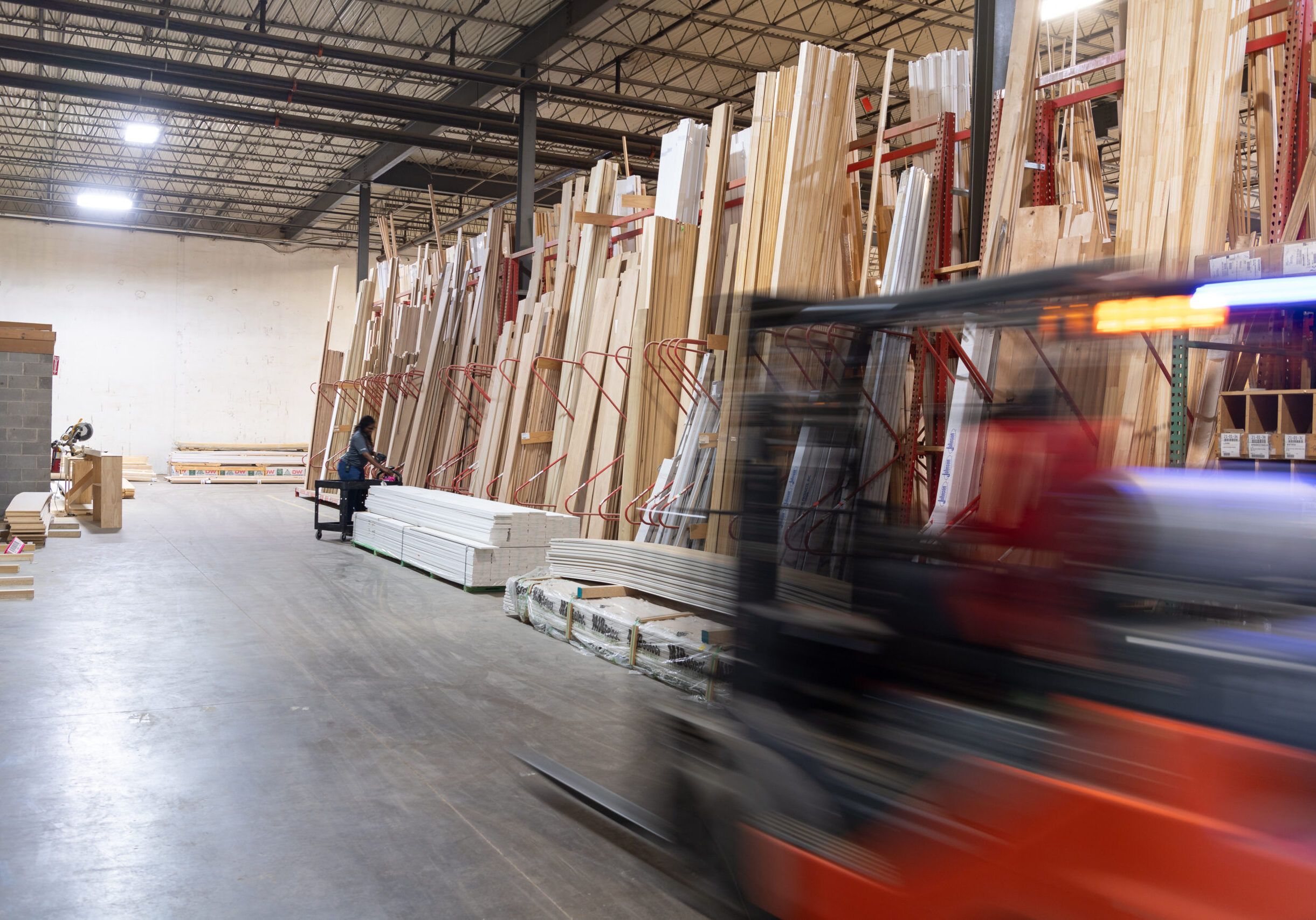 A warehouse with stacks of wood, a person using a cart, and a moving forklift.