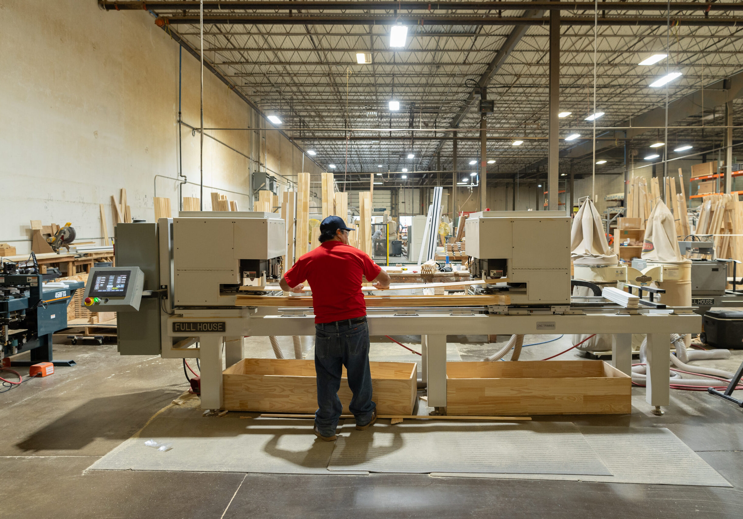KB6_5547-KevinBrownPhoto-Select A man in a red shirt and blue pants wearing a baseball cap operates automatic milling machines in a large warehouse.