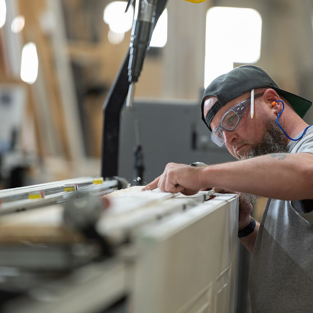 A man wearing safety glasses and a cap using a machine in a millwork workshop.